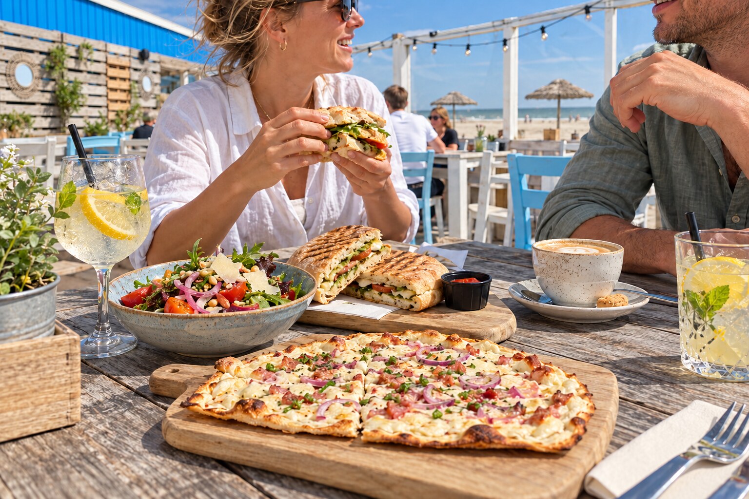 Ciabatta’s, flammkuchen en salade op een zonnige terrastafel aan zee